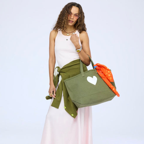 Woman wearing a pink tank and skirt with a jacket tied at the waist, carrying an olive green heart tote bag with an orange heart bandana. Shop womens accessories at Kerri Rosenthal.