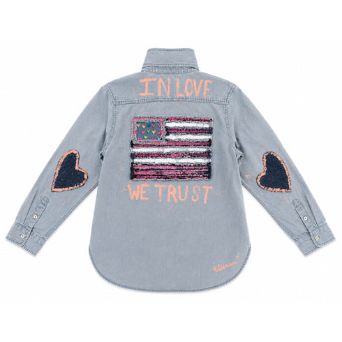 Denim shirt hand-painted on the back with 'In Love We Trust' text framing a fringed American flag patch with pink heart accents, and heart-shaped elbow patches on both sleeves — Kerri Rosenthal wearable art