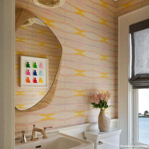 Powder room with Kerri Rosenthal pastel ikat wallpaper, a curved organic-shaped travertine mirror, a framed grid of colorful Buddha figurines, brass faucet fixtures, and a pink floral arrangement on the vanity, with a water view through the window.