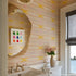 Powder room with Kerri Rosenthal pastel ikat wallpaper, a curved organic-shaped travertine mirror, a framed grid of colorful Buddha figurines, brass faucet fixtures, and a pink floral arrangement on the vanity, with a water view through the window.
