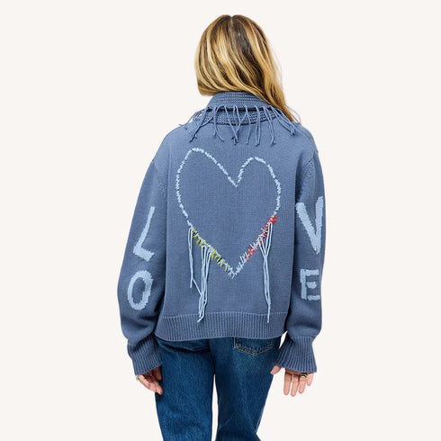Back view of a woman wearing a Kerri Rosenthal slate blue knit cardigan featuring a large intarsia heart, colorful fringe detail, and 'LOVE' lettering on the sleeves, styled with blue jeans.