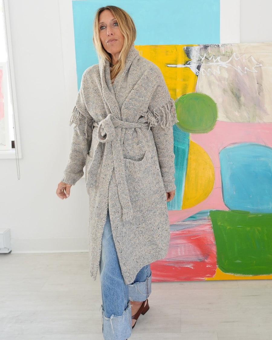 Model wearing a long gray fringe cardigan belted at the waist, cuffed jeans.