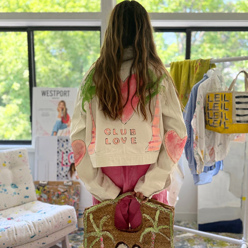Back view of a model wearing a cream cropped jacket hand-painted with a pink heart, palm trees, and 'Club Love' lettering, with pink heart elbow patches. Handpainted by Kerri Rosenthal