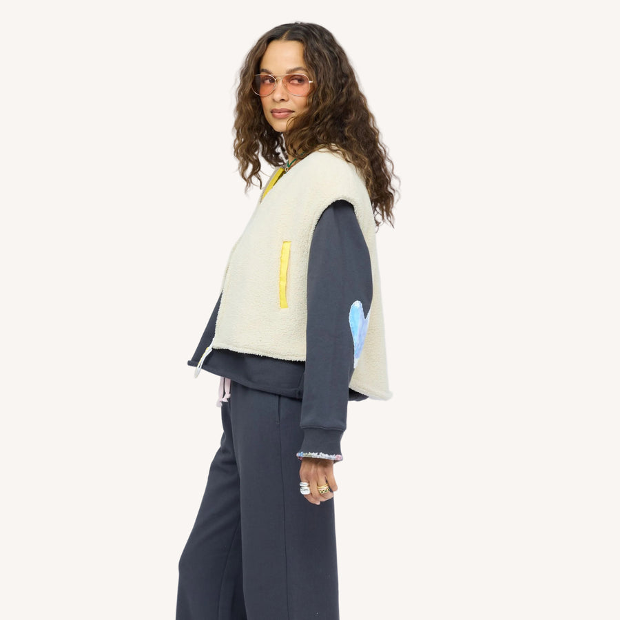 Woman wearing a cream sherpa vest with colorful patchwork sleeves over a navy sweatshirt and nave sweatpants, with sunglasses.