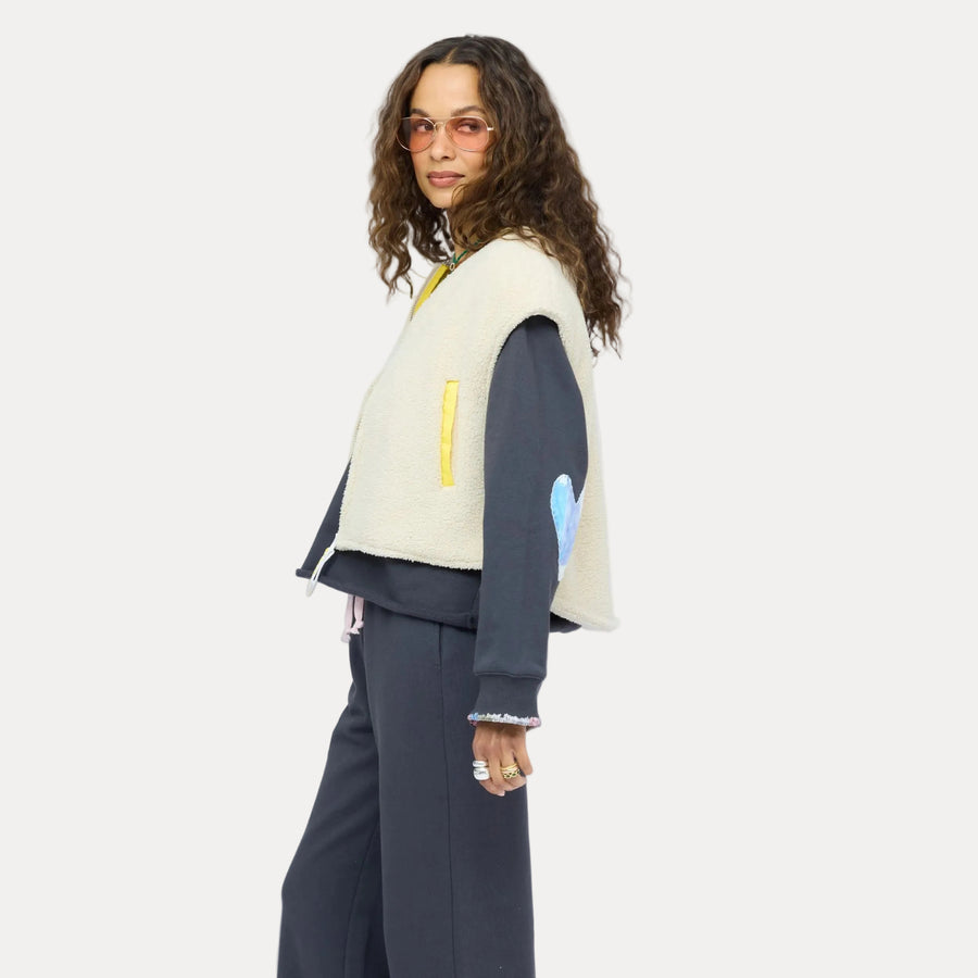 Woman wearing a cream sherpa vest with colorful patchwork sleeves over a navy sweatshirt and nave sweatpants, with sunglasses.