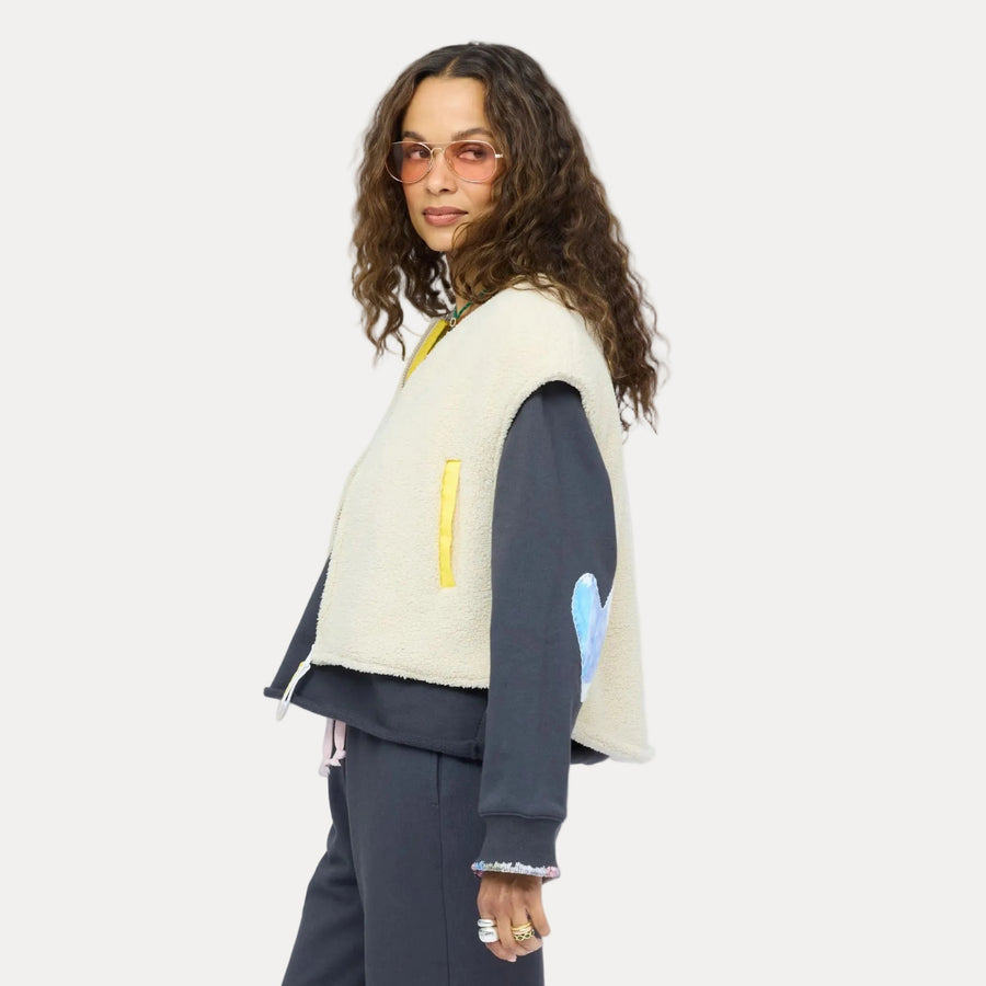 Woman wearing a cream sherpa vest with colorful patchwork sleeves over a navy sweatshirt and nave sweatpants, with sunglasses.