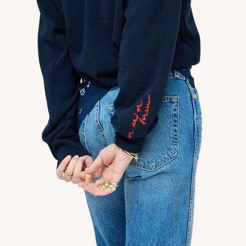 Close-up of a woman wearing a Kerri Rosenthal navy cardigan with red 'now and forever' embroidery on the sleeve, styled with blue jeans and a navy heart print bandana.