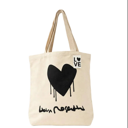 A cream-colored canvas tote bag with a black dripping heart graphic and the word 'LOVE' printed in a white square on the upper right side. The bag has two beige shoulder straps and a black script graphic underneath the heart.