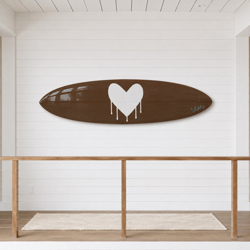 A brown surfboard-shaped decorative plaque or tray featuring a large white dripping heart in the center and a small white script signature on the right side, shown against a plain white background.