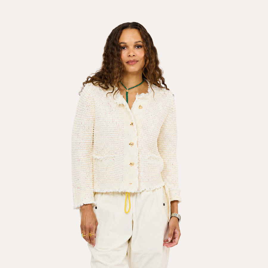 Woman wearing a Kerri Rosenthal cream boucle tweed fringe jacket with gold heart buttons, styled with white drawstring pants and a green beaded necklace.