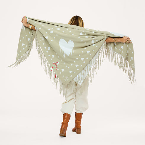 Back view of a woman holding up a sage green fringed poncho with a heart pattern and red 'love' stitching, wearing cream pants and brown boots.