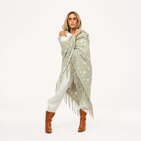 Woman wrapped in a sage green fringed poncho with a heart pattern, wearing cream pants, brown boots, and sunglasses.