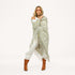 Woman wrapped in a sage green fringed poncho with a heart pattern, wearing cream pants, brown boots, and sunglasses.