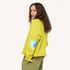 Side back view of a woman wearing a chartreuse sweatshirt with a blue heart elbow patch with paint splats and olive pants.