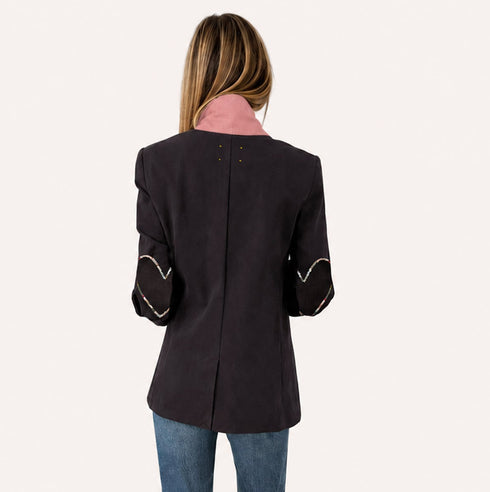 Back view of a woman wearing a coal blazer with heart embroidery on the sleeves, a pink collar, and jeans.