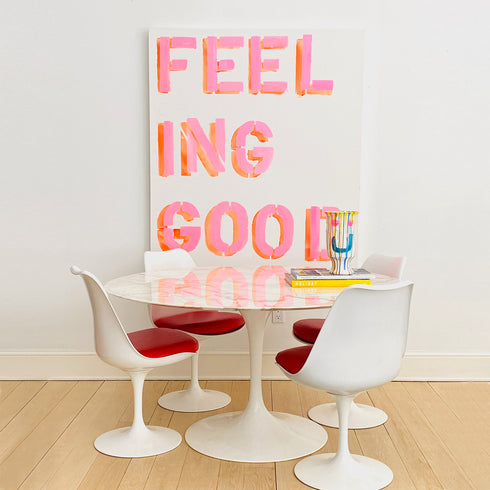 A modern dining area featuring a round white table with tulip-style white chairs that have red seats, set against a white wall with a large colorful art print that reads 'FEELING GOOD'.