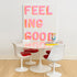 A modern dining area featuring a round white table with tulip-style white chairs that have red seats, set against a white wall with a large colorful art print that reads 'FEELING GOOD'.