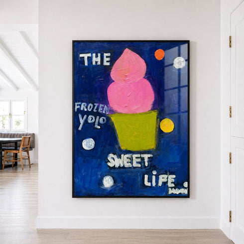 A framed square art print hanging on a white wall in a bright room. The artwork features a pink frozen yogurt on a dark blue background with white text reading 'THE FROZEN YOLO SWEET LIFE.