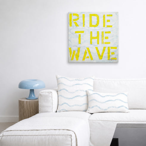 Large rectangular artwork featuring the words Ride the Wave in blue and yellow by Kerri Rosenthal, hanging on a neutral wall above a modern white sectional sofa styled with matching blue patterned pillows.