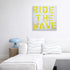 Large rectangular artwork featuring the words Ride the Wave in blue and yellow by Kerri Rosenthal, hanging on a neutral wall above a modern white sectional sofa styled with matching blue patterned pillows.