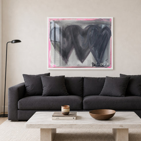 Holding Hearts art print by Kerri Rosenthal with three black hearts and grey ground and pink outline in a white frame displayed on a living room wall with black couch.