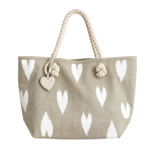 A canvas tote bag in a light taupe-beige color with a white ikat heart pattern, featuring thick cream-colored rope handles and a small heart-shaped charm.