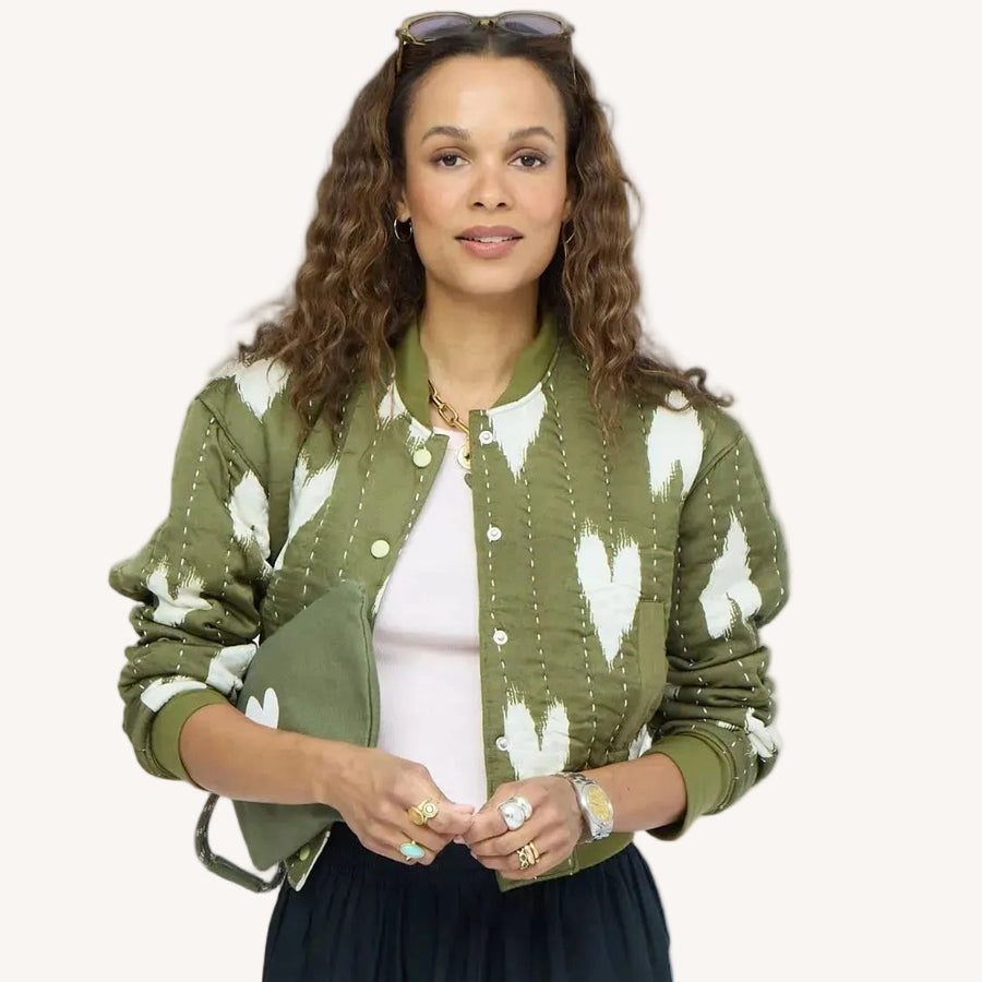 Woman wearing an olive green quilted bomber jacket with heart print, white top, and sunglasses on her head.