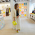 Kerri wearing a cream crochet Frankie cardigan with neon yellow-green trim, a white flowy maxi kr two tone tank dress and lime green wedge sandals, holding a chartreuse heart clutch bag indoors.