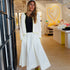 Kerri standing in the Westport flagship shop wearing a white open-knit crochet cardigan with neon yellow-green trim over a black tank and white flowy maxi skirt with lime green sandals, colorful drippy heart painting visible in the background.