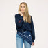 Close-up of woman wearing a Kerri Rosenthal navy v-neck cardigan with red 'now and forever' sleeve embroidery and a navy Kerri Rosenthal heart and smiley print bandana tied at the waist, styled with blue jeans and a gold chain necklace."