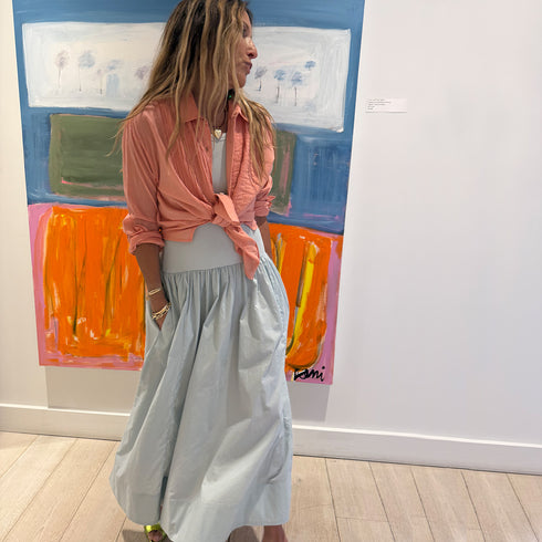 Kerri standing in her Westport gallery wearing a powder blue gathered maxi KR Tank dress and a coral pink tied button-down Tuxedo shirt, styled with gold jewelry and green heeled sandals in front of a colorful abstract painting.