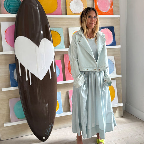 Kerri standing beside a chocolate brown drippy heart surfboard in an art gallery wearing a pale blue denim jacket and matching gathered maxi dress with lime green sandals, colorful circle paintings displayed on shelves behind her.