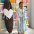 Kerri standing beside a chocolate brown drippy heart surfboard in an art gallery wearing a pale blue denim jacket and matching gathered maxi dress with lime green sandals, colorful circle paintings displayed on shelves behind her.