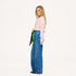 Side view of a woman wearing a Kerri Rosenthal pink and cream striped long sleeve crewneck top with a gold heart detail, styled with wide-leg blue jeans, a green and navy heart print bandana, and pink sunglasses.