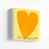 Love Looks Good on You art print: a bold orange heart with an angular shape on a pale yellow background, with "Love Looks Good On You" hand-painted below it in black, printed on stretched canvas