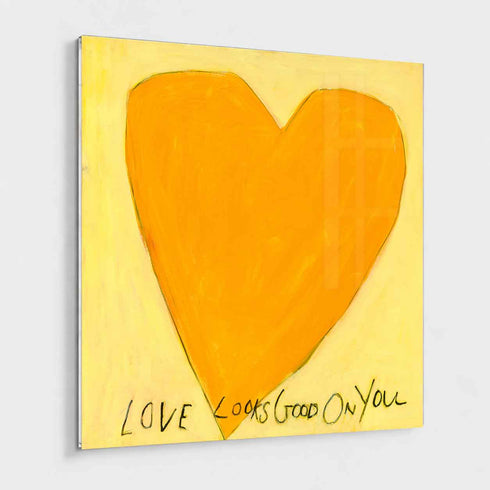 Love Looks Good on You art print: a bold orange heart with an angular shape on a pale yellow background, with "Love Looks Good On You" hand-painted below it in black, mounted under plexi with a frameless finish