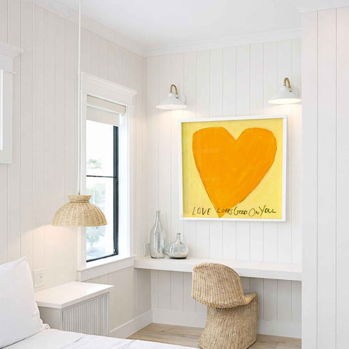 Love Looks Good on You art print: a bold orange heart with an angular shape on a pale yellow background, with "Love Looks Good On You" hand-painted below it in black, hung in a room setting