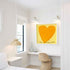 Love Looks Good on You art print: a bold orange heart with an angular shape on a pale yellow background, with "Love Looks Good On You" hand-painted below it in black, hung in a room setting