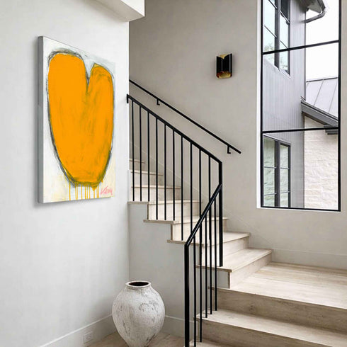 Love Note 1 art print: a bold orange heart with a rounded, tulip-like shape and a charcoal outline with paint drips, hung on a wall beside a staircase in an entryway with a large ceramic vase below it