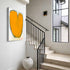 Love Note 1 art print: a bold orange heart with a rounded, tulip-like shape and a charcoal outline with paint drips, hung on a wall beside a staircase in an entryway with a large ceramic vase below it