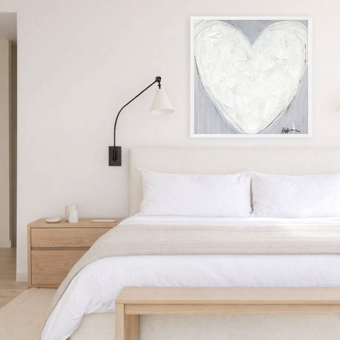 Morning My Love art print, a white heart painting in a white frame, hung above a bed with white linens in a neutral, light-wood bedroom