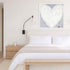 Morning My Love art print, a white heart painting in a white frame, hung above a bed with white linens in a neutral, light-wood bedroom