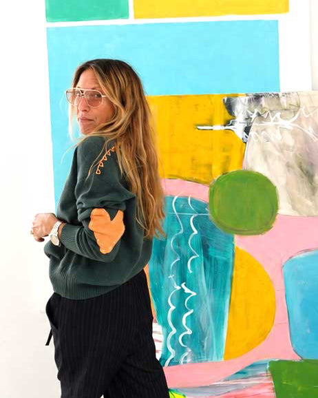 Kerri in a green heart-patch sweater and black pinstripe pants standing next to a colorful abstract painting.