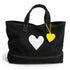 A black canvas tote bag with white contrast stitching, featuring a large white heart on the front and a small yellow heart-shaped tag hanging from a yellow string.