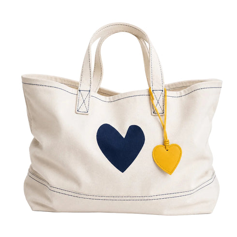 An off-white canvas tote bag with a navy blue heart on the front and a yellow leather heart charm hanging from the handle, set against a plain white background.