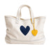 An off-white canvas tote bag with a navy blue heart on the front and a yellow leather heart charm hanging from the handle, set against a plain white background.