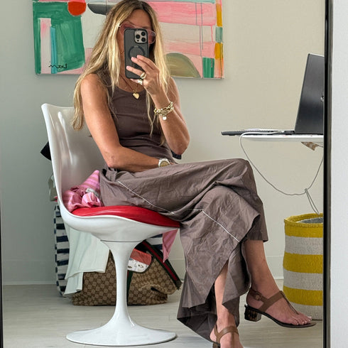 Mirror selfie of a woman seated in a white and red tulip chair, wearing a Kerri Rosenthal brown tank dress with contrast white piping, gold jewelry, and brown thong sandals. Colorful abstract painting hangs on the wall behind her.