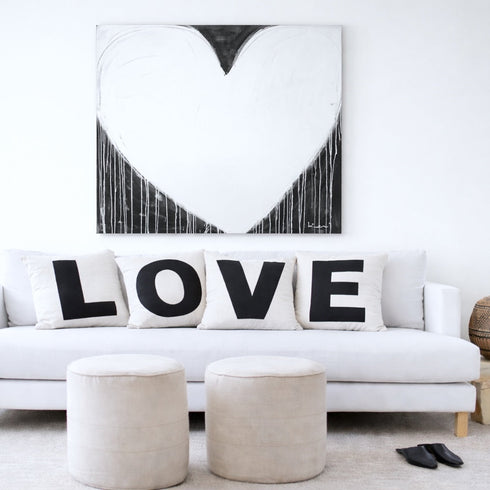White sofa with 'LOVE' cushions, two ottomans, and a heart-shaped abstract painting in a living room.