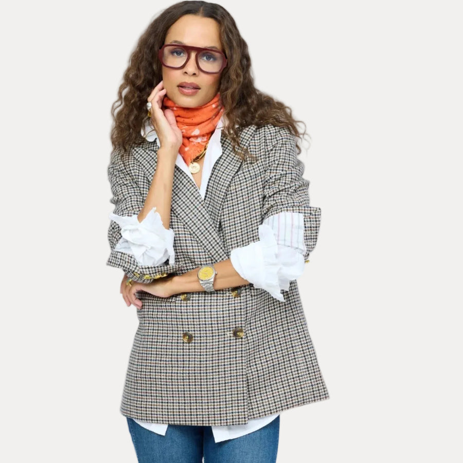 A woman wearing a double-breasted houndstooth blazer in beige, brown, and black, styled with a white shirt, orange scarf, and blue jeans, set against a solid light gray background.