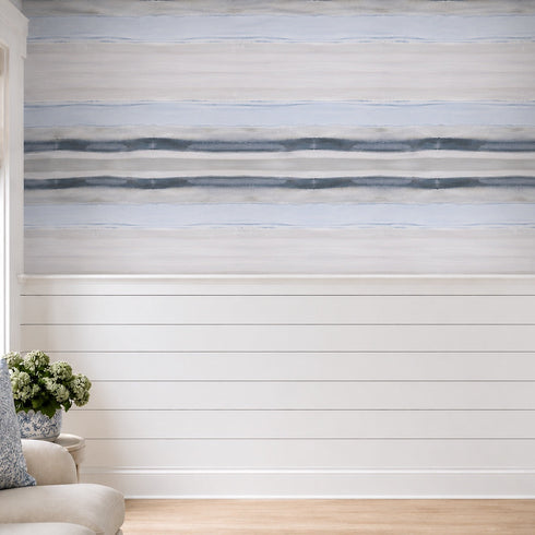 A wall mural or wallpaper featuring a horizontal beach-inspired design with light blue and white textured stripes above a white shiplap lower wall, shown in a styled room with a sofa, pillow, and window.